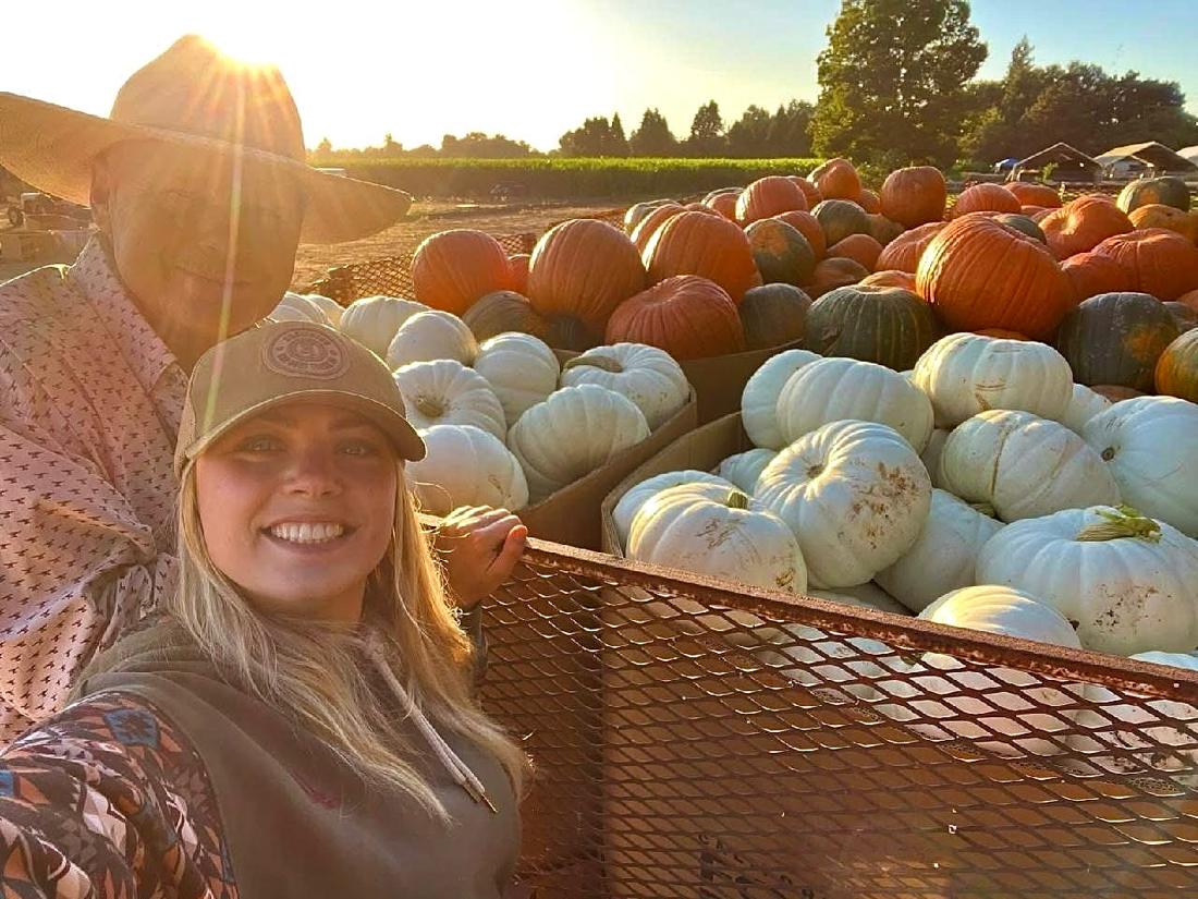 Two members of the Zittel Farms family smiling beside a trailer loaded with white and orange pumpkins at sunset